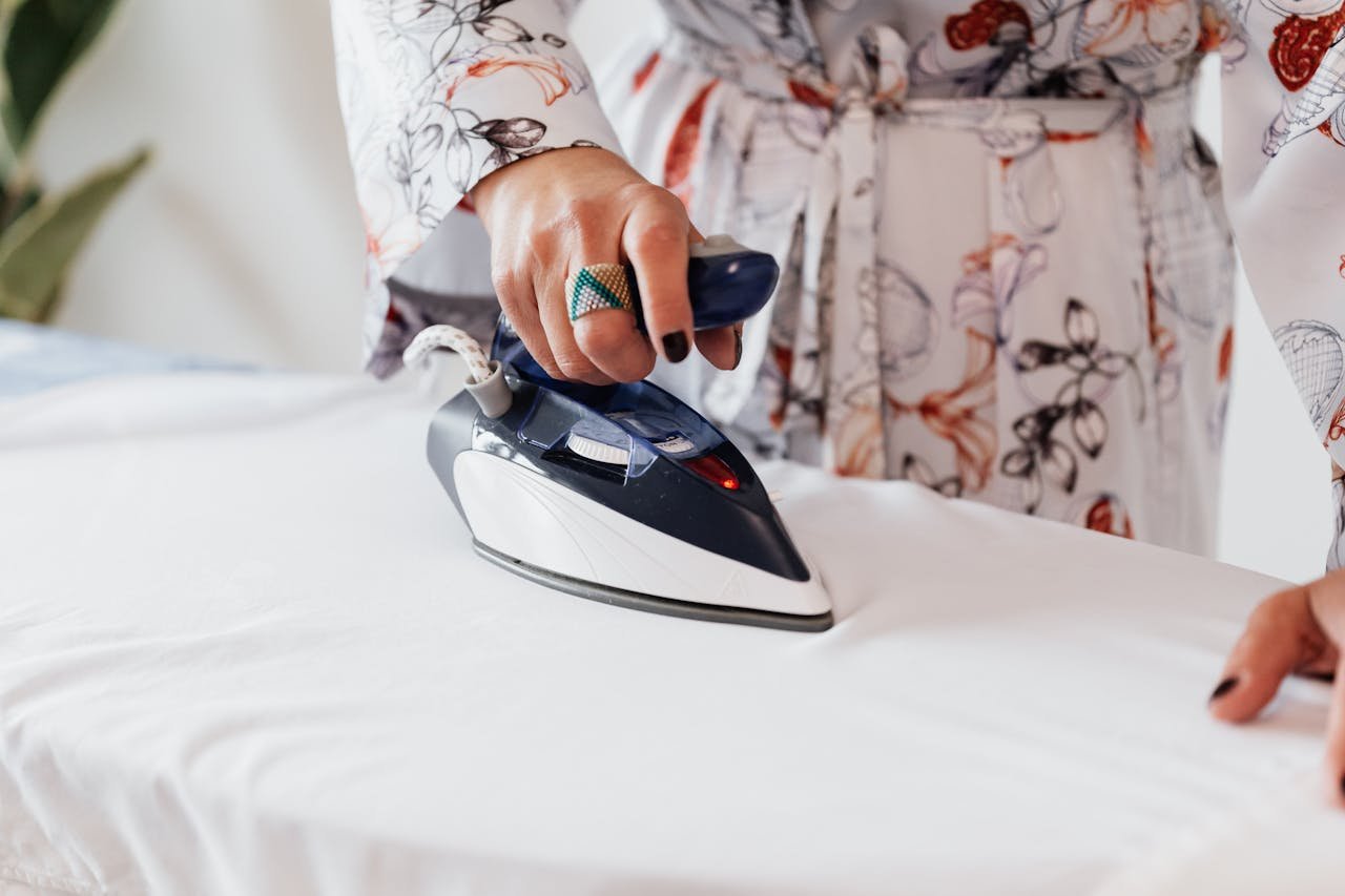 Crop unrecognizable female with manicured hands wearing classy silk robe and stylish ring ironing white sheets on ironing board